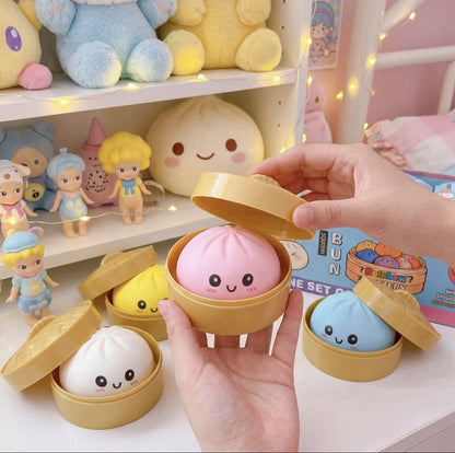 A set of cute, round buns in small bamboo-style containers, with a hand lifting the lid of the central pink bun. Other buns in white, yellow, and blue are visible on a white shelf in a decorated room with plush toys and fairy lights.